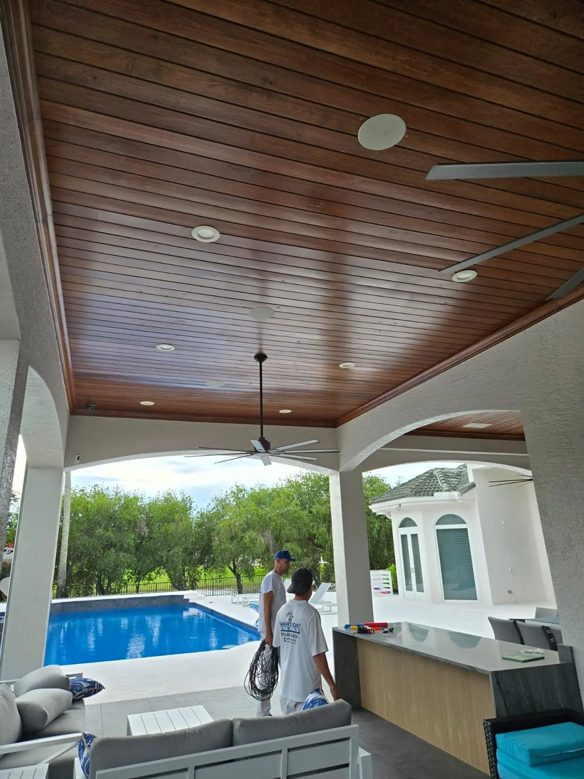 Two painters on a patio under a freshly finished wood-grain ceiling, a pool beyond