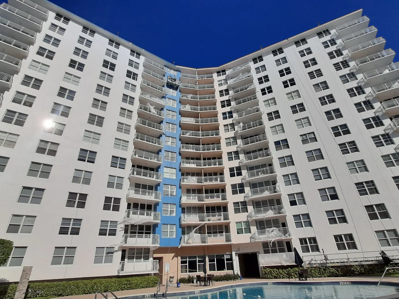 White high-rise condominium with a blue-accented center tower above a pool and palms