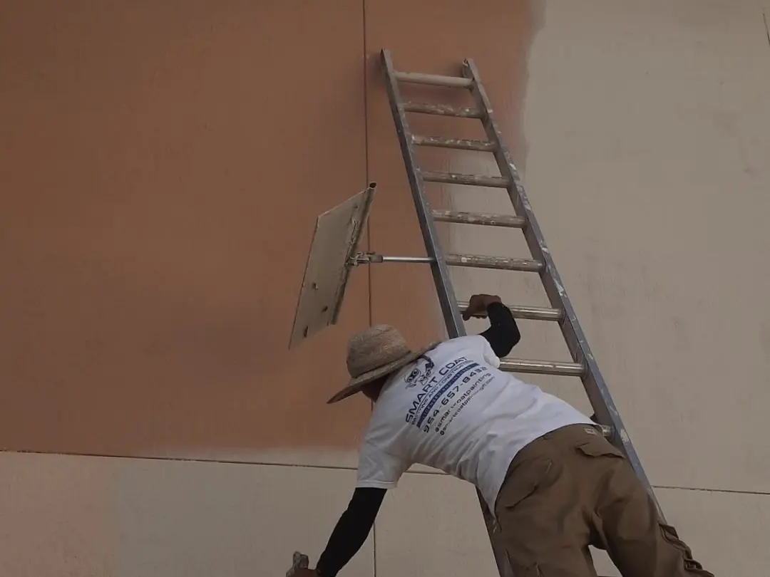 Painter on a ladder spraying a two-tone stucco wall, the new coat going on over the old