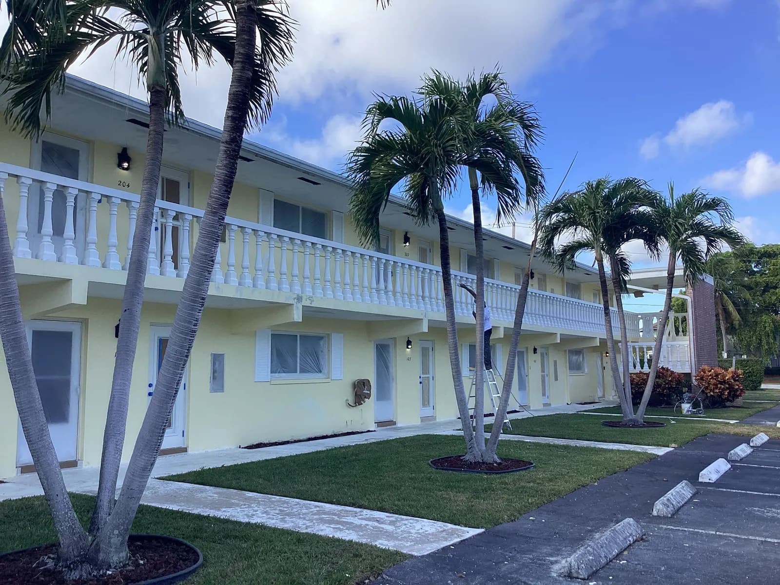 Two-story garden apartments repainted pale yellow with white balustrades behind palms