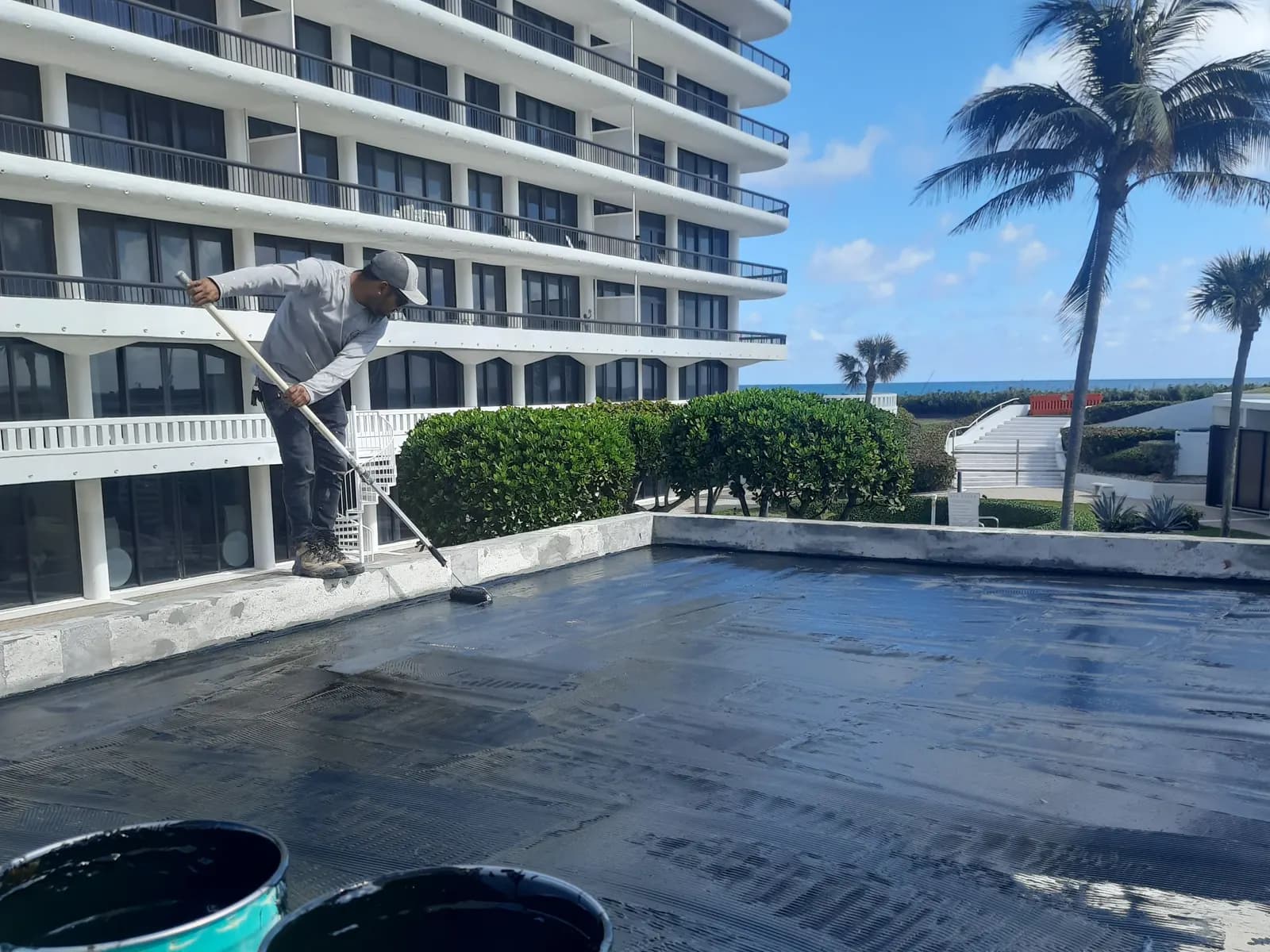 Crew rolling a black waterproofing membrane onto a plaza deck beside an oceanfront high-rise